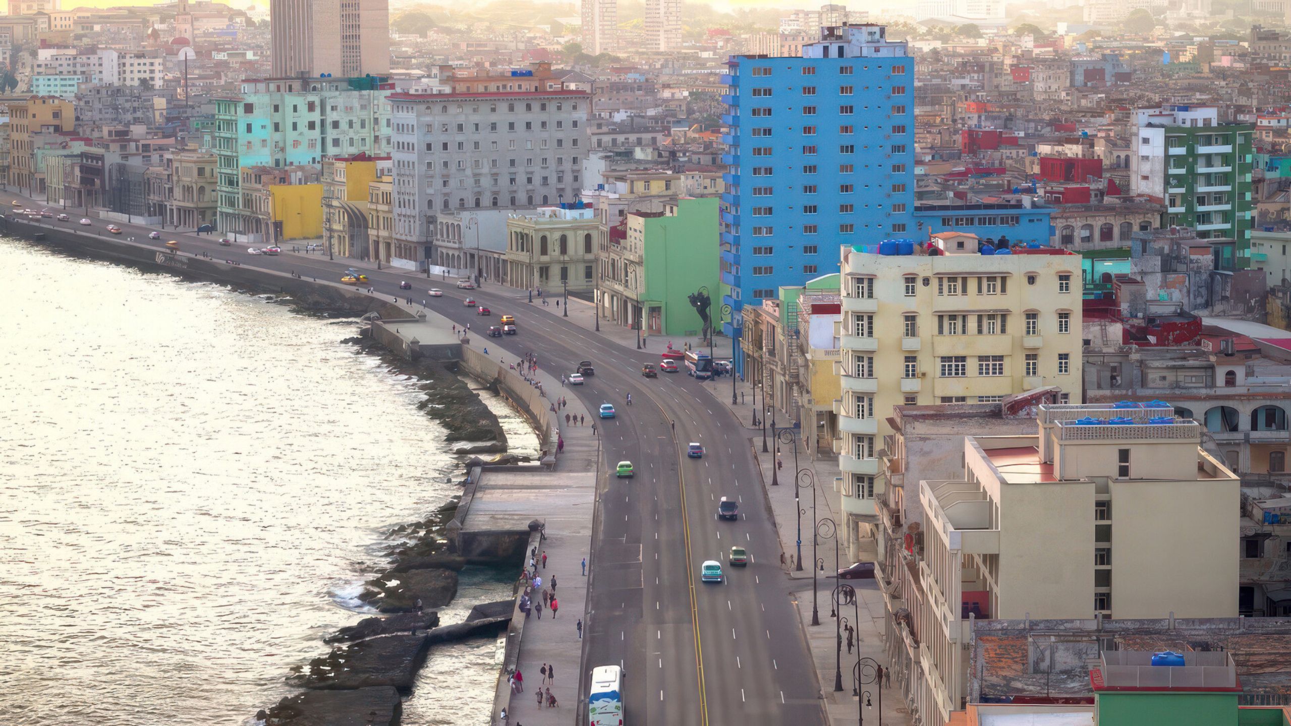 A road runs close to the Havana coastline. Colorful blue, green, and yellow cars and buildings dot the city.