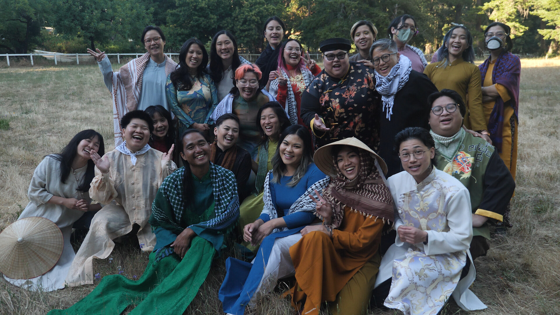 A group of queer trans Vietnamese members all huddled up (standing and sitting) wearing a variety of traditional Vietnamese garments called 