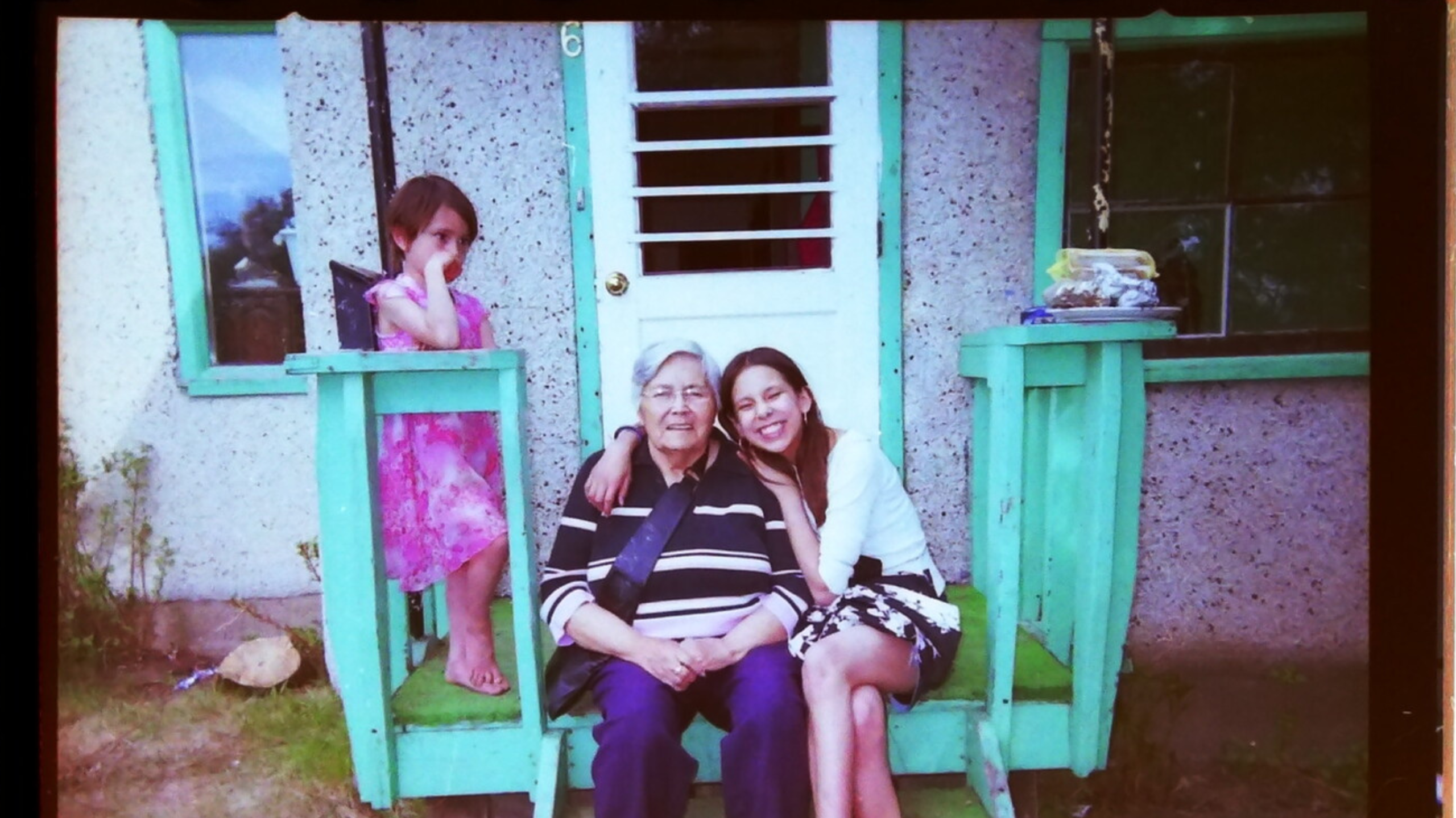 A still for IndigiQueer Stewards. A faded photo of three people sitting on the front steps of a small house with a turquoise-painted door and bench. An elderly person with gray hair sits in the center wearing a black and white striped shirt, with a young woman in a white top and floral skirt smiling and leaning against them on the right. A small child in a pink dress stands on the bench to the left, partially covering their face with one hand.