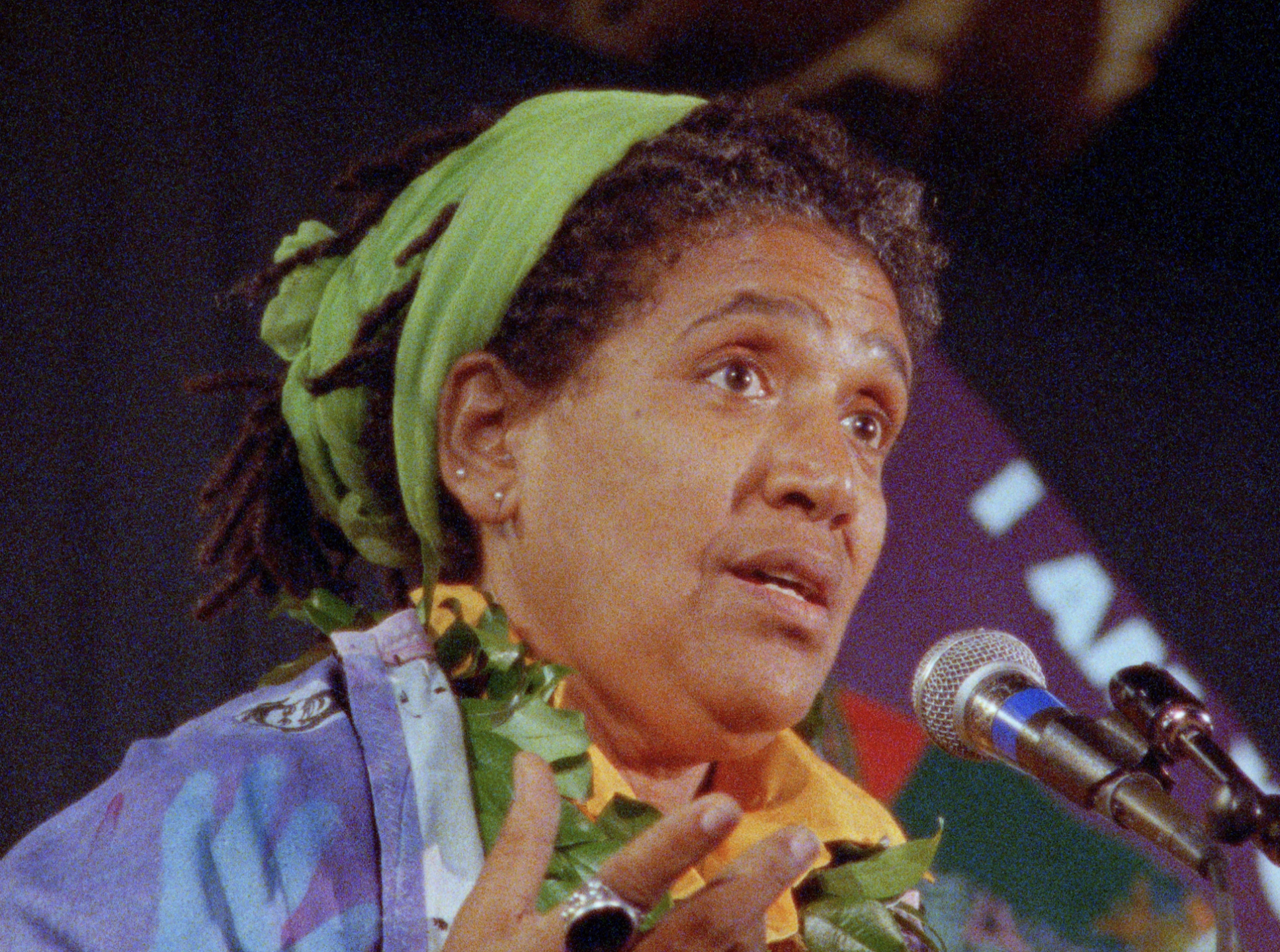 A still from A Litany for Survival: The Life and Work of Audre Lorde by Ada Gay Griffin & Michelle Parkerson.  A close-up portrait of Audre Lorde speaking at a microphone. She wears a bright green head wrap, gold hoop earrings, a yellow collared shirt under a pastel patterned jacket, and a green leaf lei around her neck.
